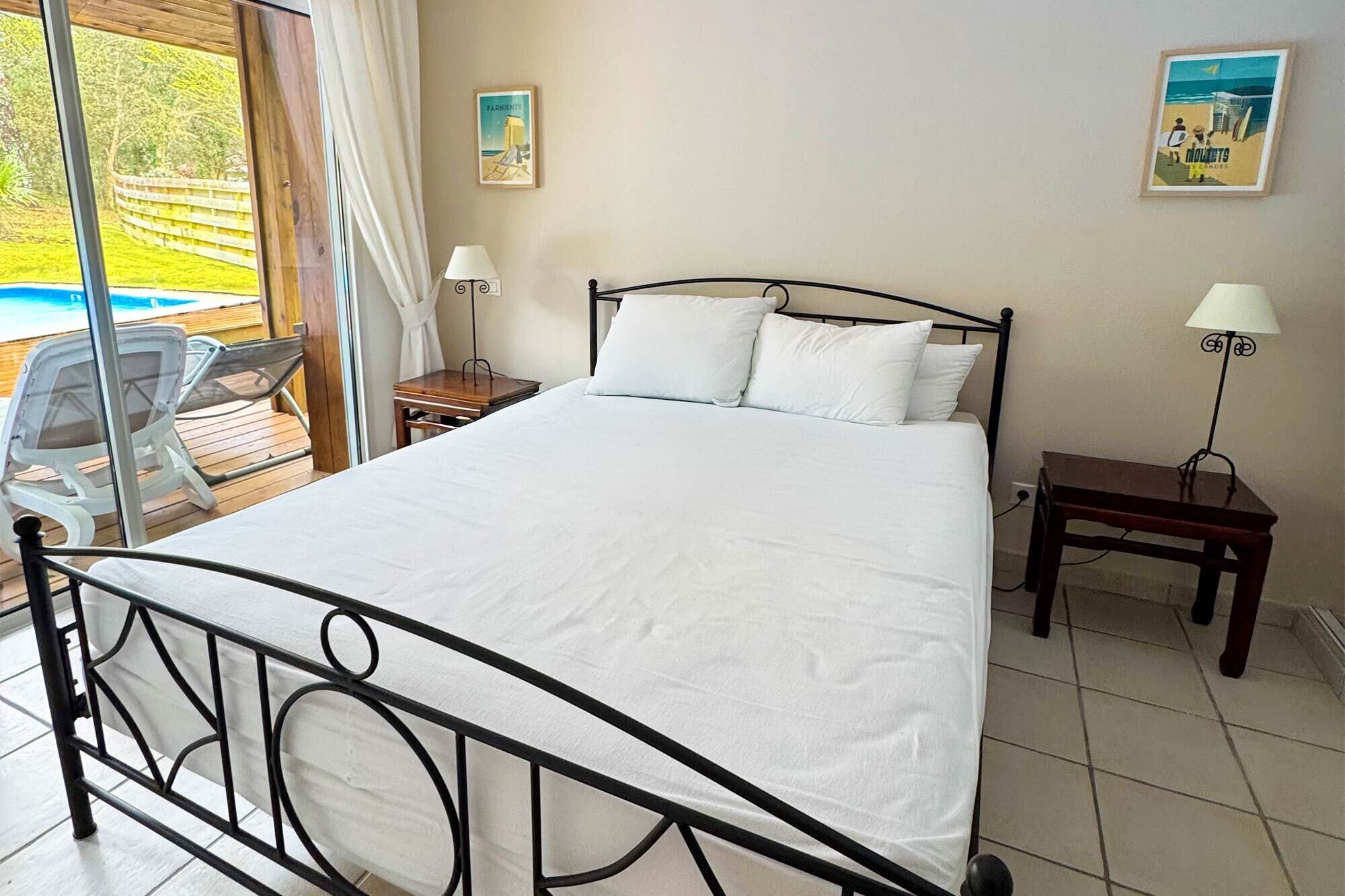 A bedroom with a bed, two bedside tables, a chair, and a view of a pool.