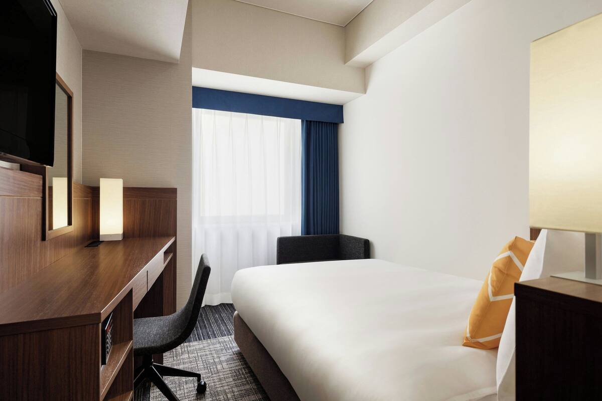A hotel room with a bed, a desk, a chair, a TV, and a window with curtains.