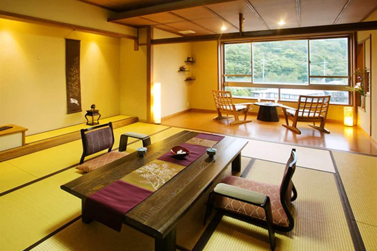 Ein traditioneller japanischer Raum mit Tatami-Böden, einem niedrigen Tisch mit Kissen und Blick auf einen Wald durch große Fenster.
