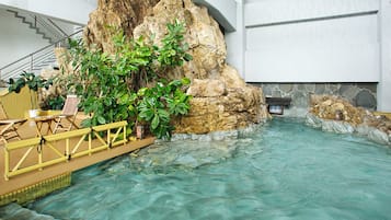 An indoor pool with clear water, a wooden deck, and a rock wall with plants.