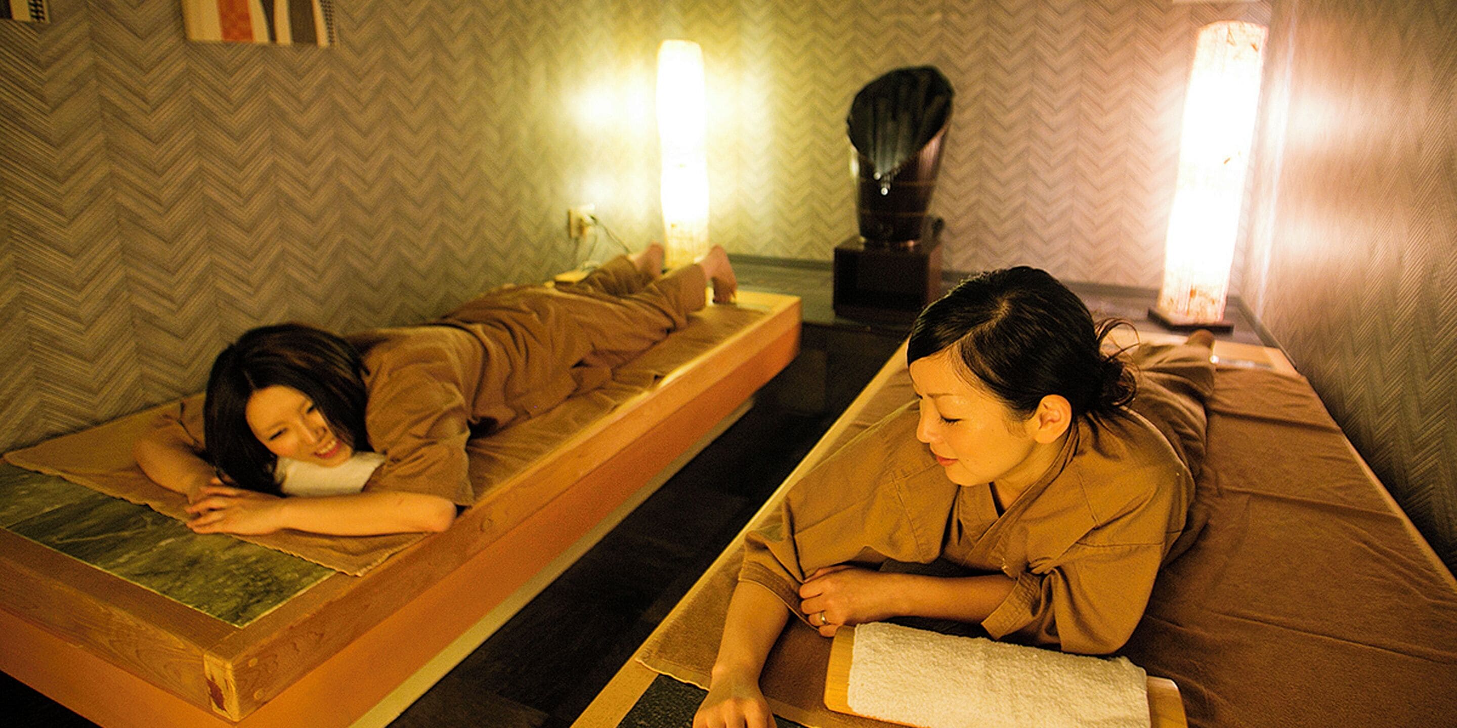 Two individuals lying on separate wooden beds in a room with a herringbone-patterned wall and two wall-mounted lamps.