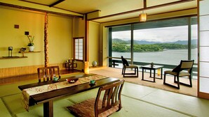 Een traditionele Japanse kamer met tatami vloeren, een lage tafel en stoelen. Een groot raam biedt uitzicht op een meer en bergen.