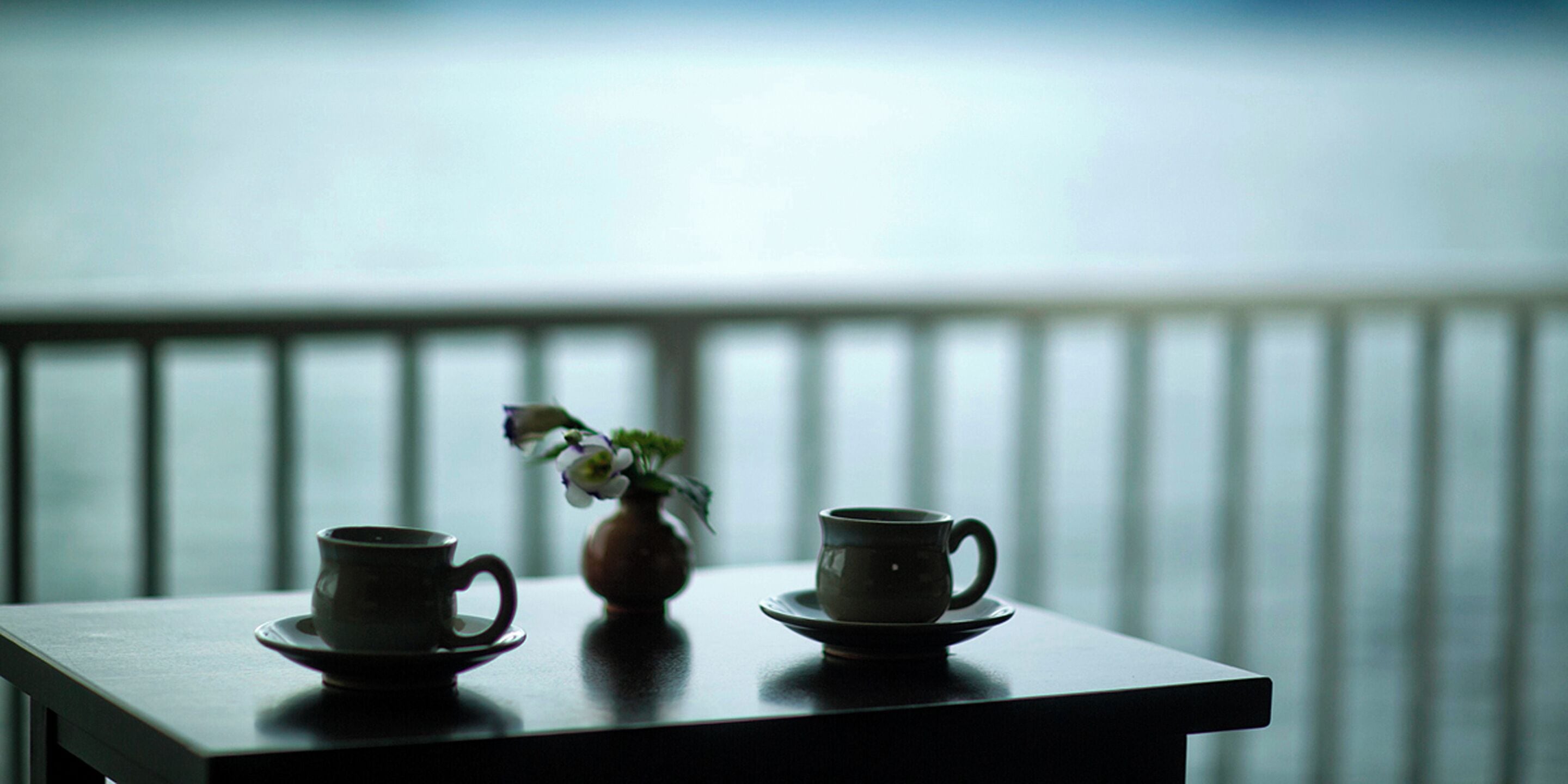 A table with two cups and a vase on a balcony overlooking a railing.