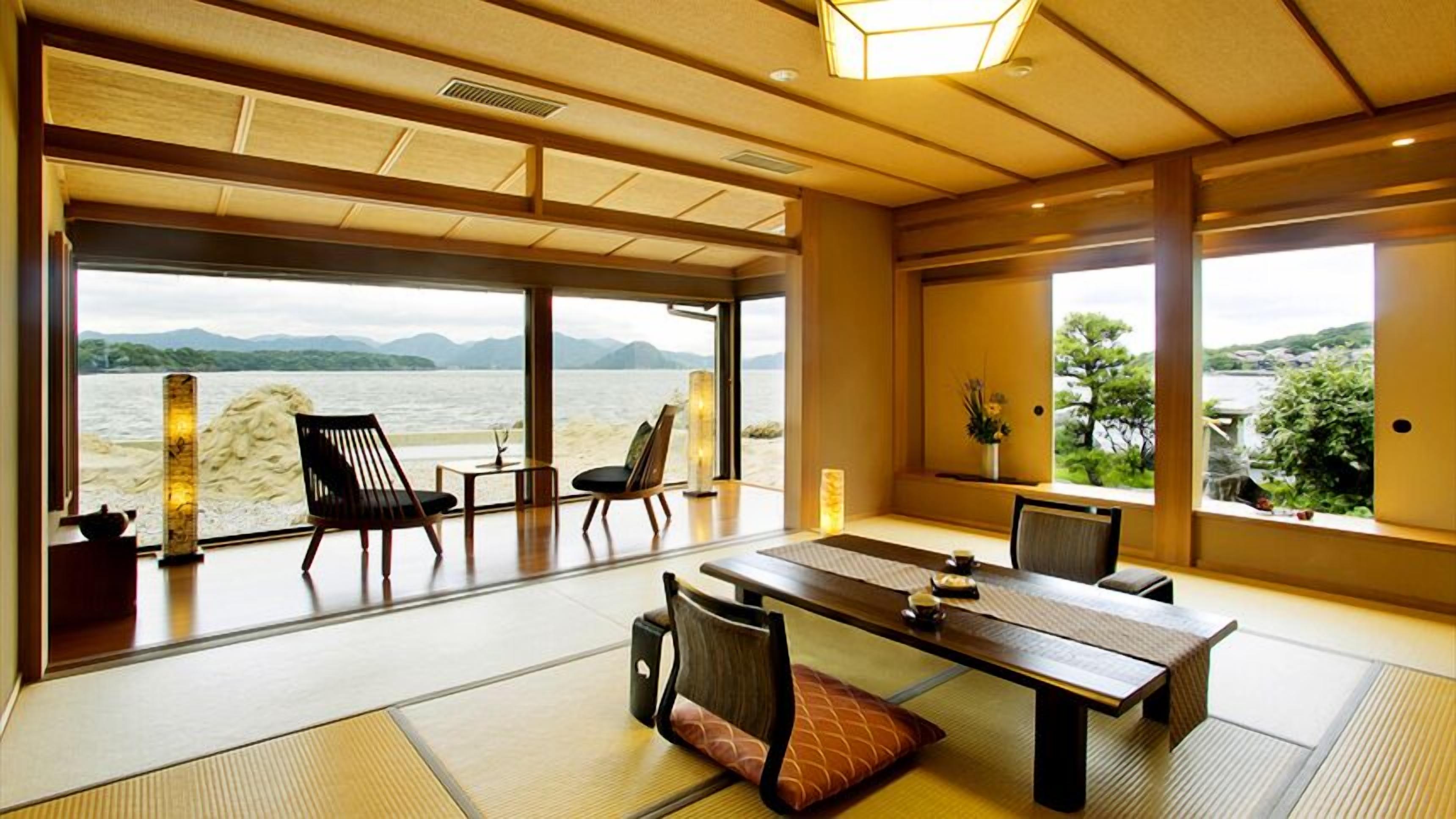 Una camera tradizionale giapponese con pavimento in tatami, un tavolino e sedie, con vista su un corpo d'acqua e montagne.