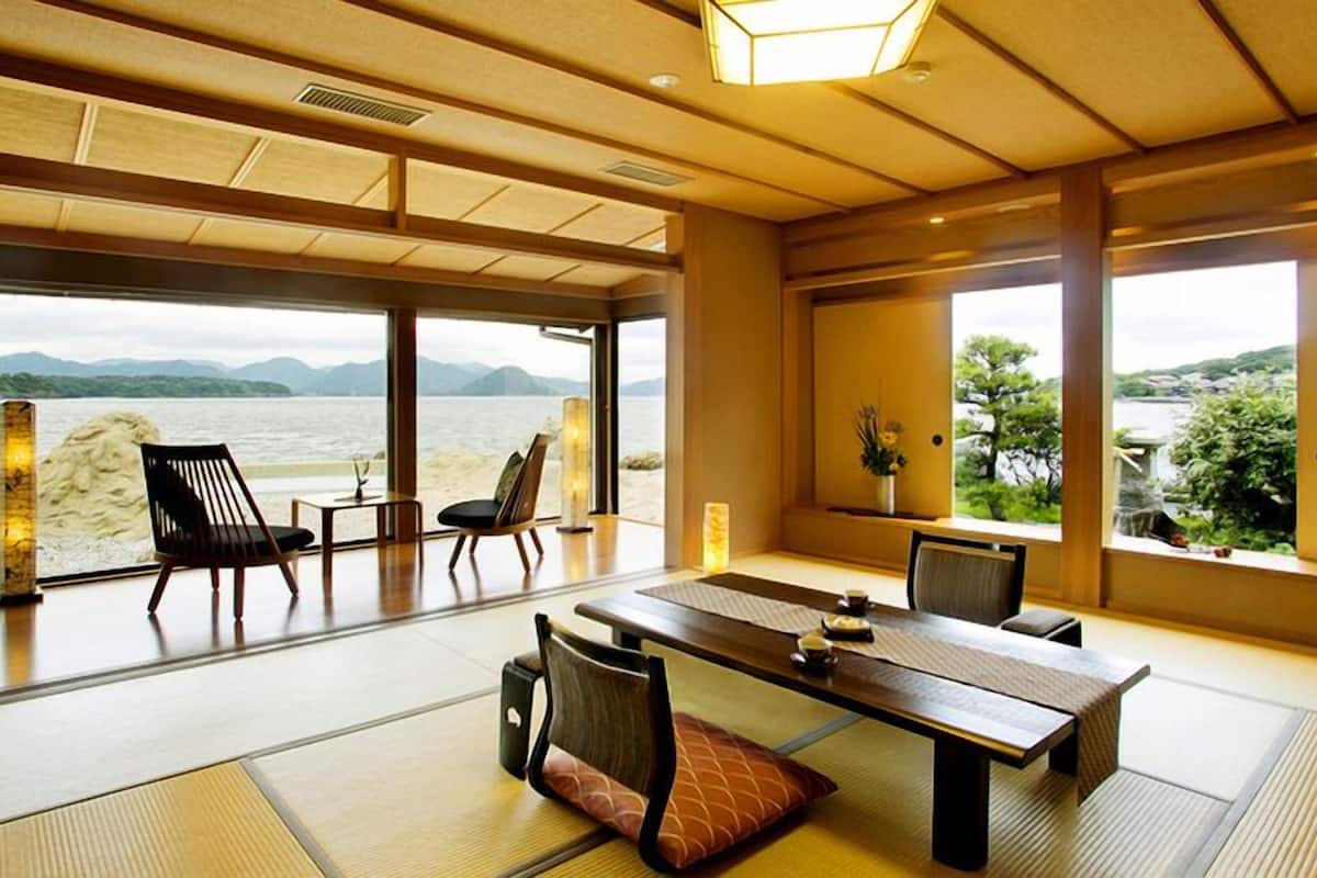 Habitación japonesa tradicional con suelo de tatami, una mesa baja y sillas, con vistas a un cuerpo de agua y montañas.