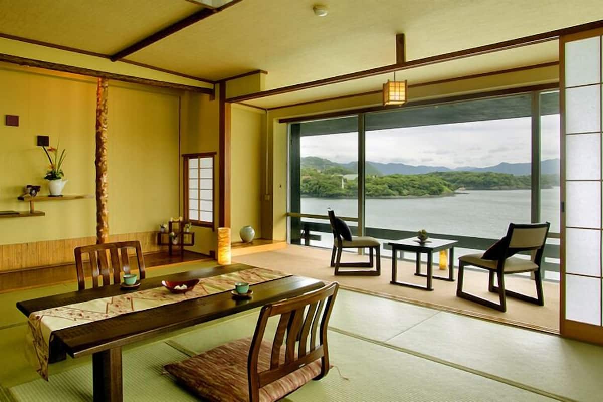 Una habitación tradicional japonesa con suelo de tatami, una mesa baja, sillas, vista a un lago y un puertas correderas.
