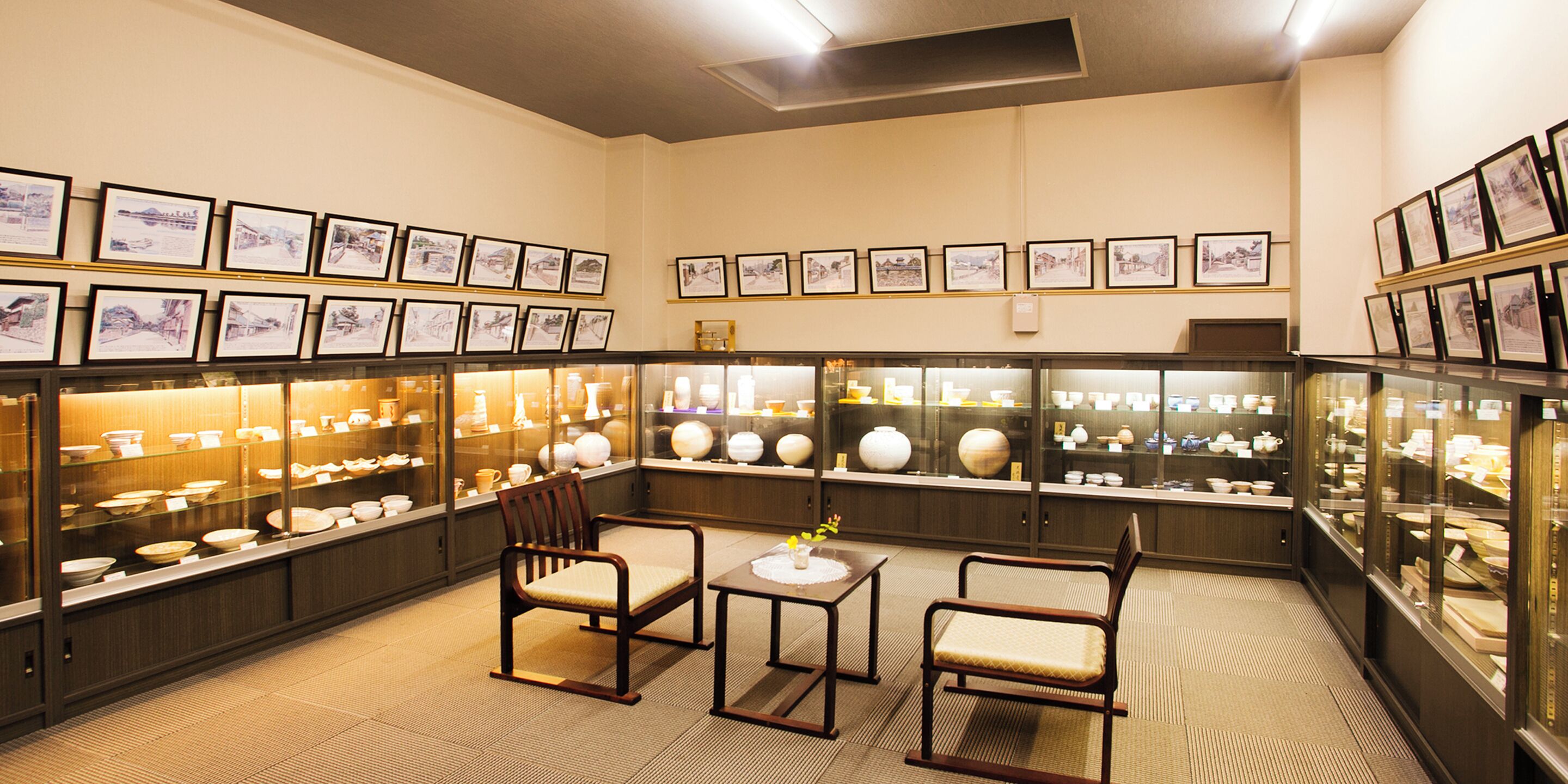 A room with a long display of pottery and ceramics, framed pictures on the walls, and a seating area with a small table.