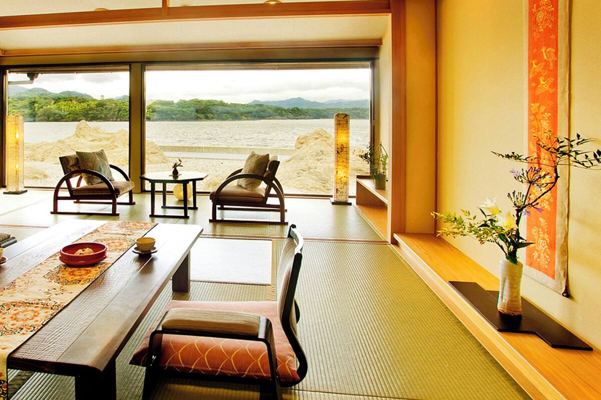 Ein traditioneller japanischer Raum mit Tatami-Bodenbelag, einem niedrigen Tisch mit Tischläufer und zwei gepolsterten Stühlen, mit Blick auf einen See durch große Fenster.