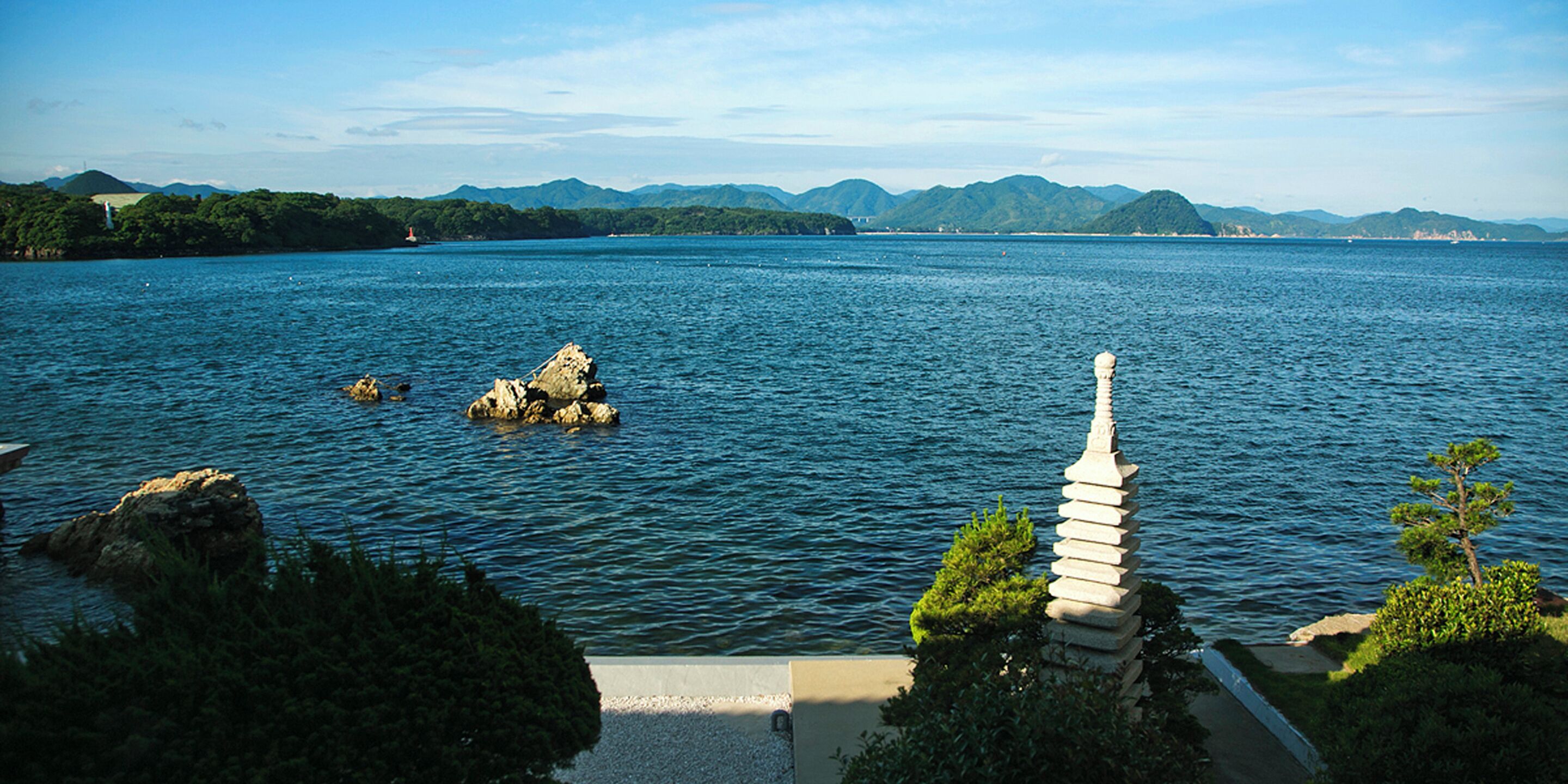 A serene lake with a pagoda and rocky outcrops, surrounded by lush greenery and distant mountains.