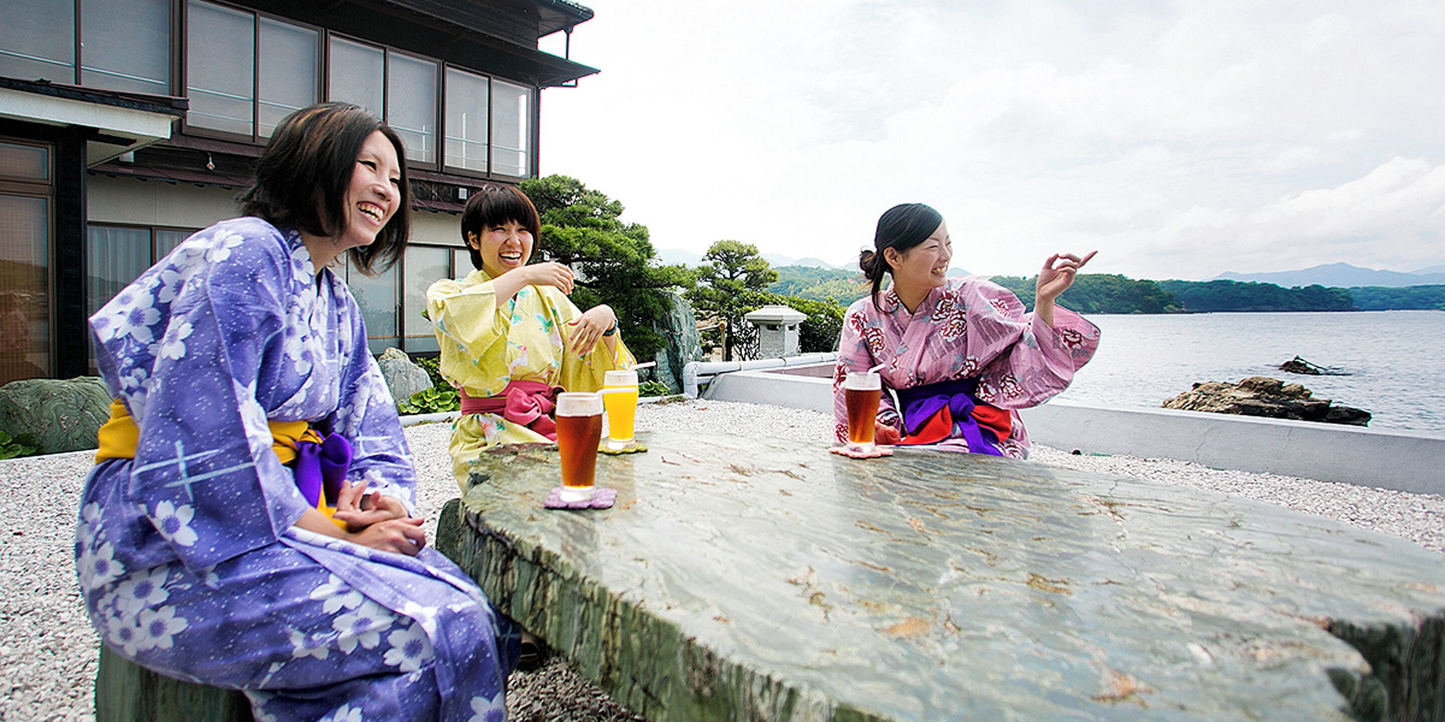 A group of people sitting at a wooden table outdoors with drinks, near a body of water and a building.