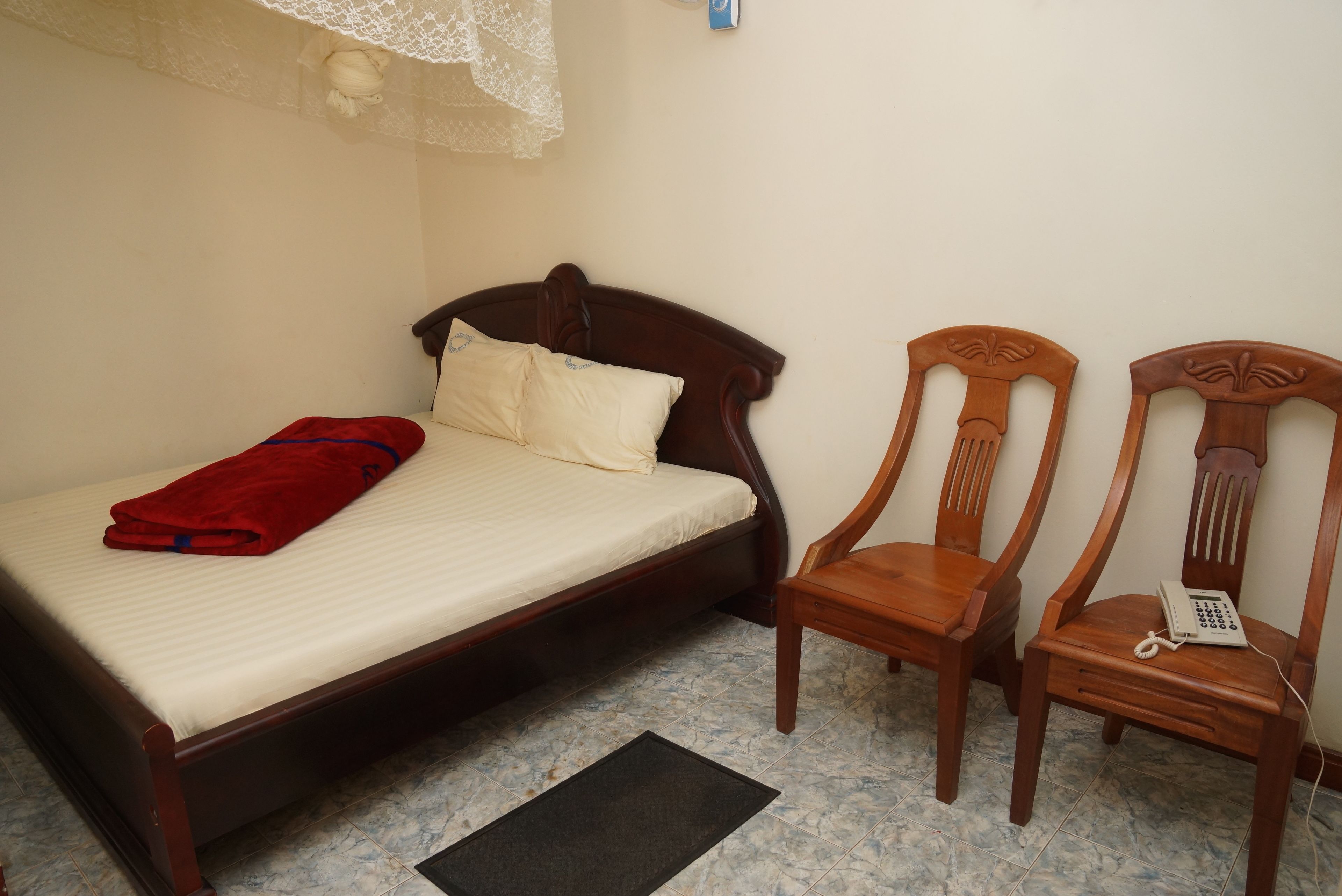 A single bed with a red blanket, two wooden chairs, and a bedside telephone.