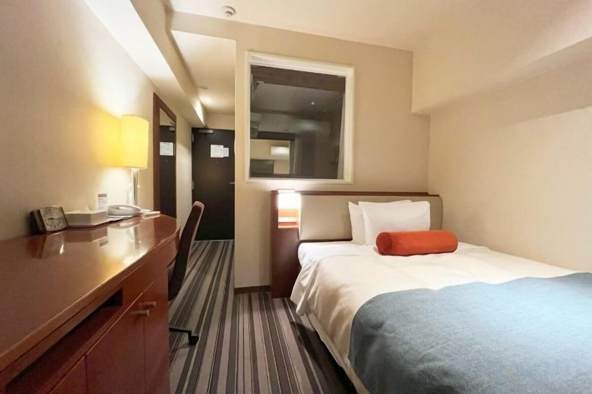Ein Hotelzimmer mit Bett, Schreibtisch, Stuhl, Lampe und Fenster.