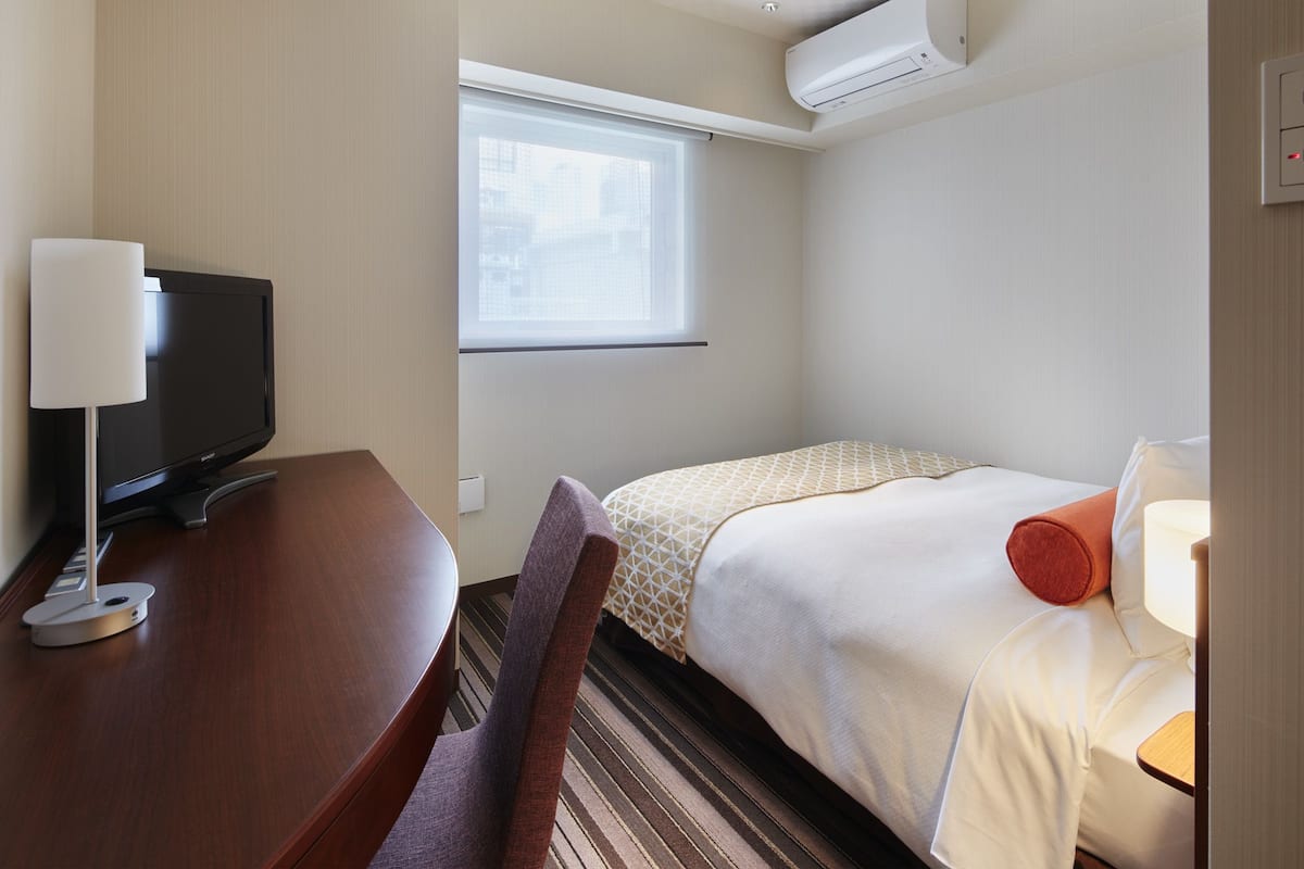 A hotel room with a bed, a desk with a TV, a chair, and a window.