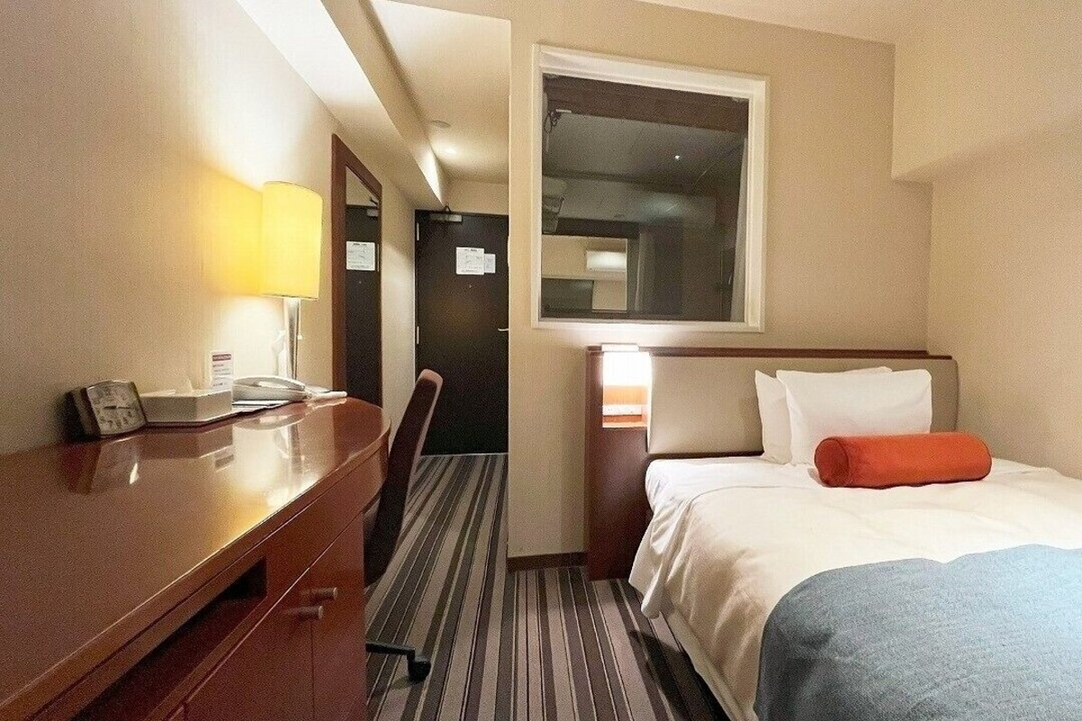Ein Hotelzimmer mit Bett, Schreibtisch mit Lampe, Stuhl und Fenster.