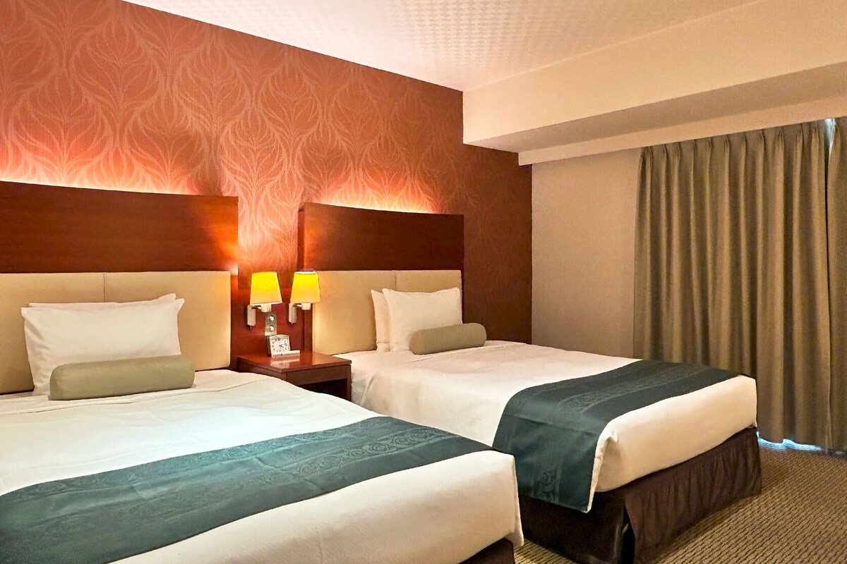 Ein Hotelzimmer mit zwei Betten, einem Kopfteil mit einer Uhr, zwei Nachttischlampen und einem großen Fenster mit Vorhängen.