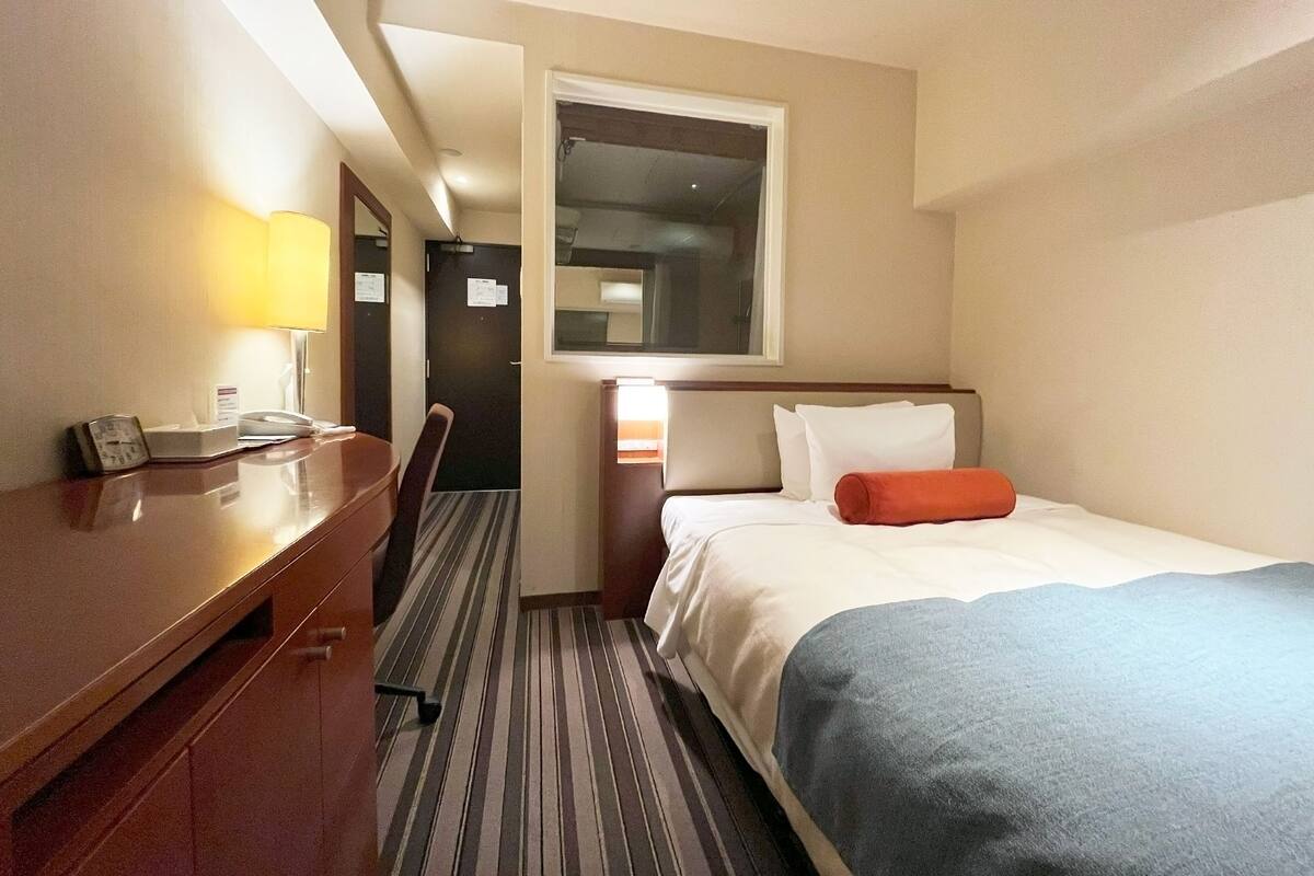 A hotel room with a bed, a desk, a chair, a lamp, and a window.