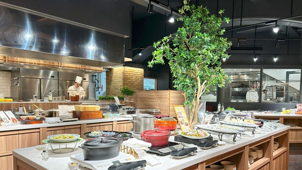 A modern kitchen with stainless steel appliances, wooden cabinets, and a buffet setup with various dishes and a large potted plant. - Nakajimaya Grand Hotel (Shizuoka)