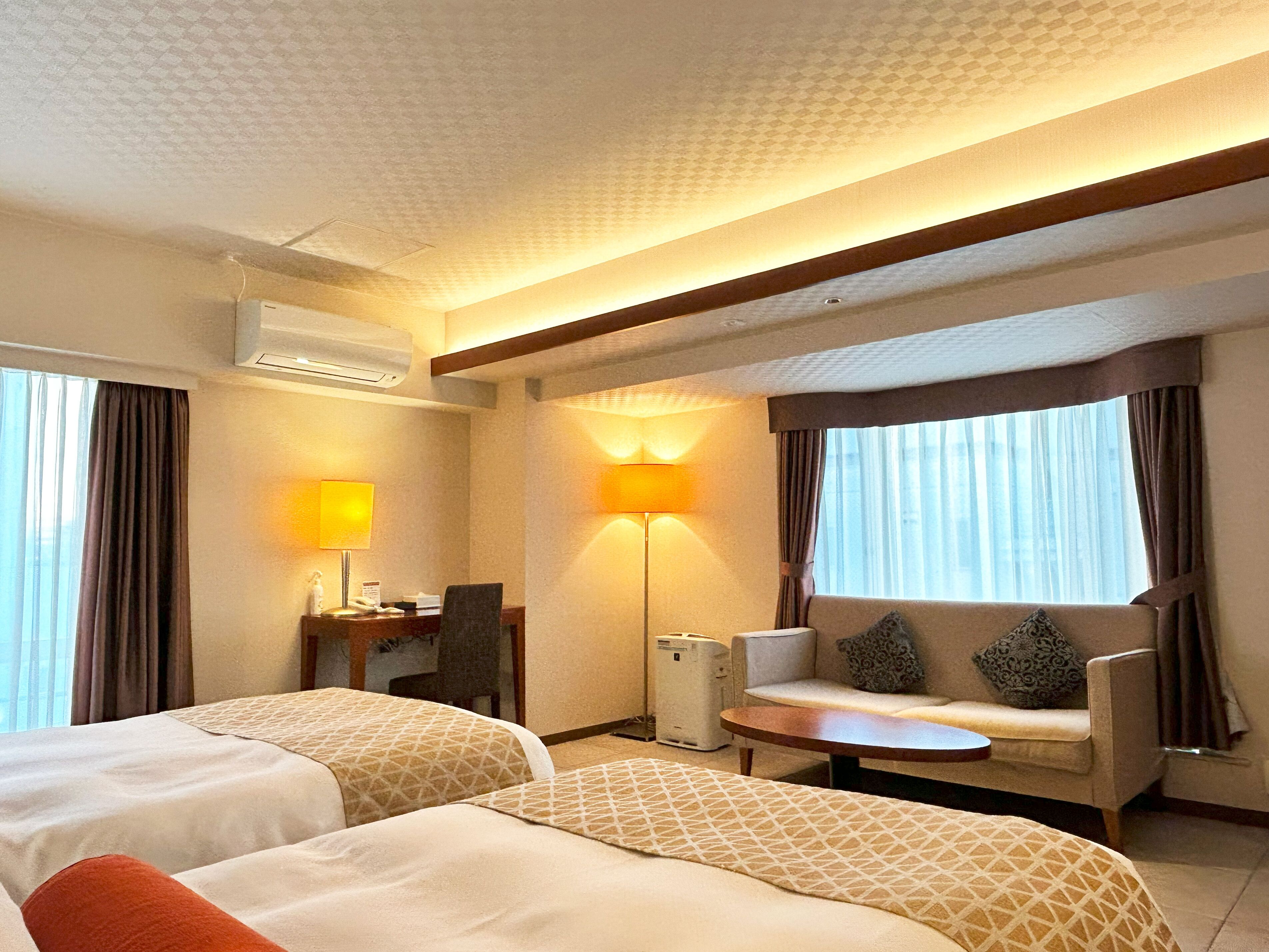 A hotel room with two beds, a desk with a lamp, a sofa, and a small table.