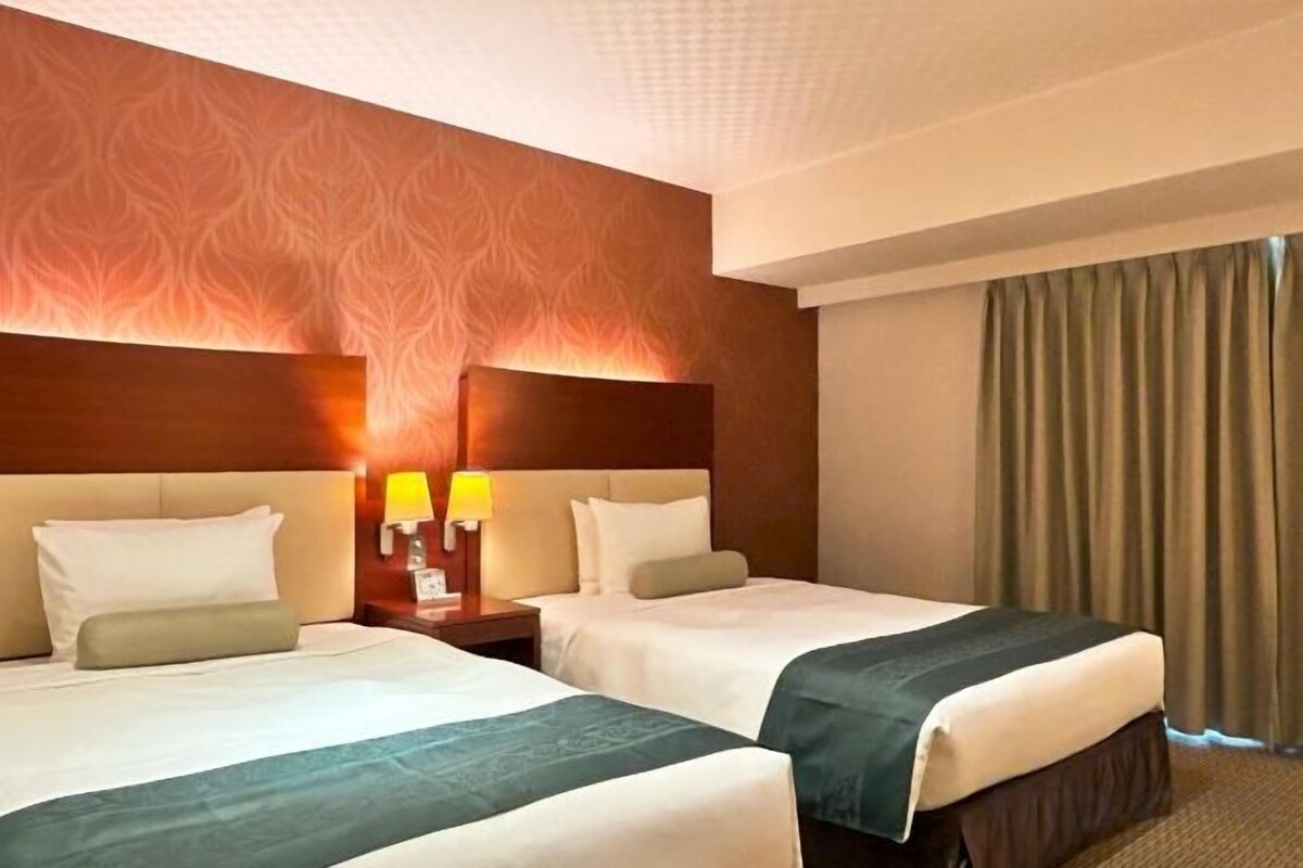 Ein Hotelzimmer mit zwei Betten, einem an der Wand befestigten Kopfteil mit einem gemusterten Design, Nachttischen mit Lampen und Blick auf einen Vorhang.