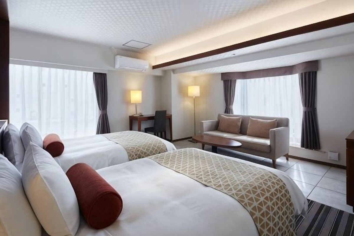 Ein Hotelzimmer mit zwei Betten, einem Schlafsofa, einem Schreibtisch und einem Fenster mit Vorhängen.