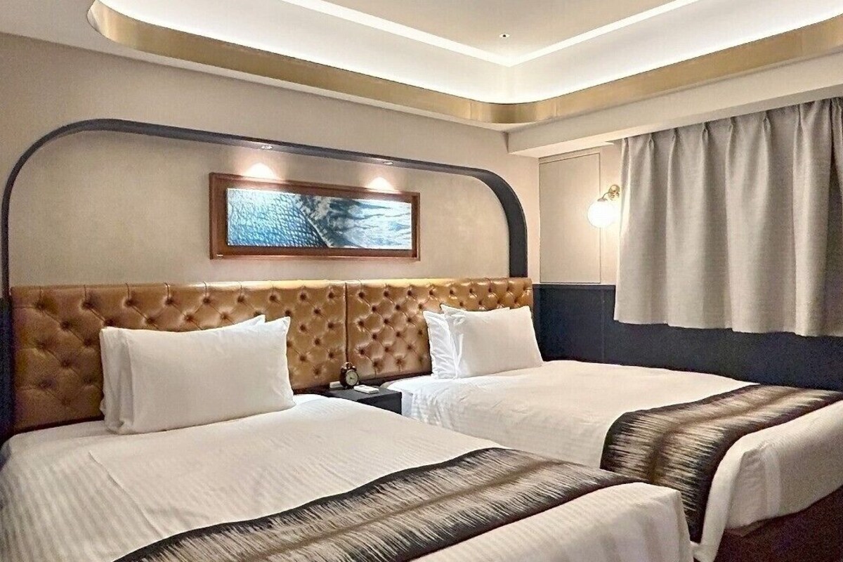 Ein Hotelzimmer mit zwei Betten, einem gepolsterten Kopfteil, einem gerahmten Kunstwerk darüber und einem Fenster mit Vorhängen.