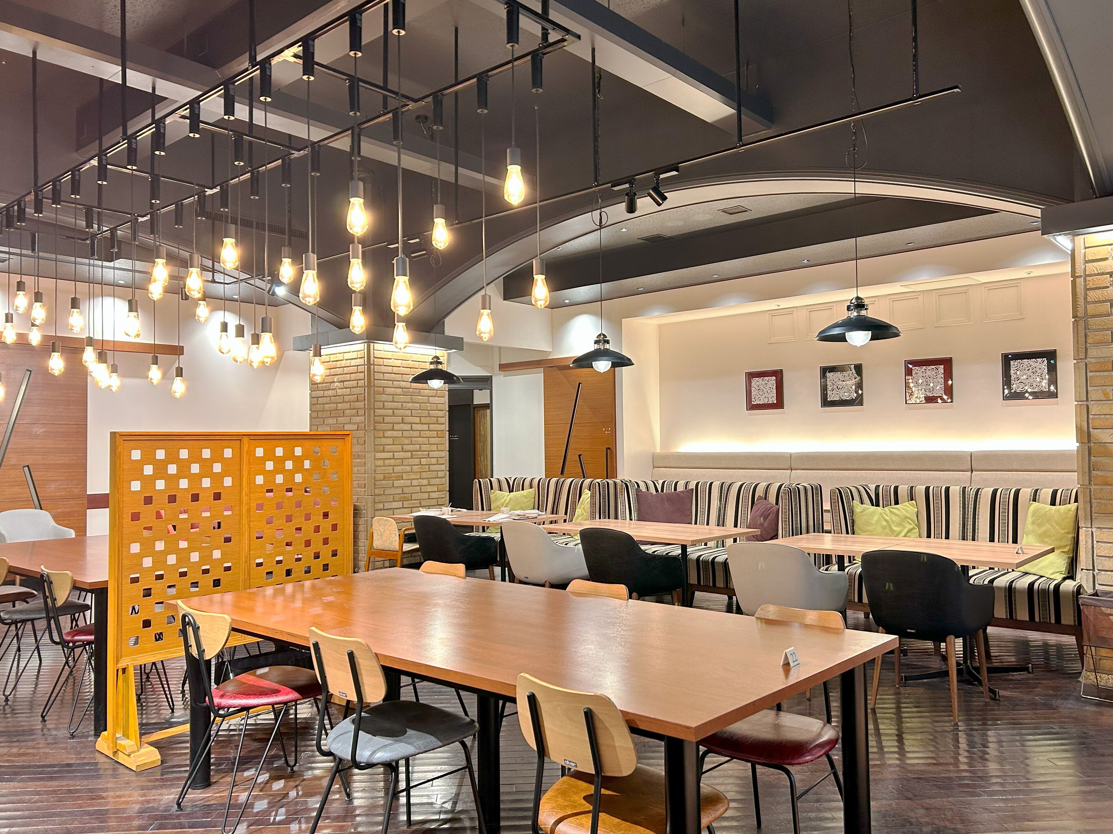 A modern restaurant with wooden tables and striped booths, hanging light fixtures, and a brick accent wall.