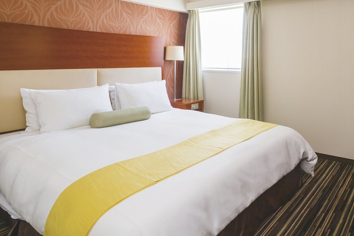 Ein Hotelzimmer mit einem großen Bett, einem Holz-Kopfteil, einem Nachttisch mit Lampe und einem Fenster mit Vorhängen.