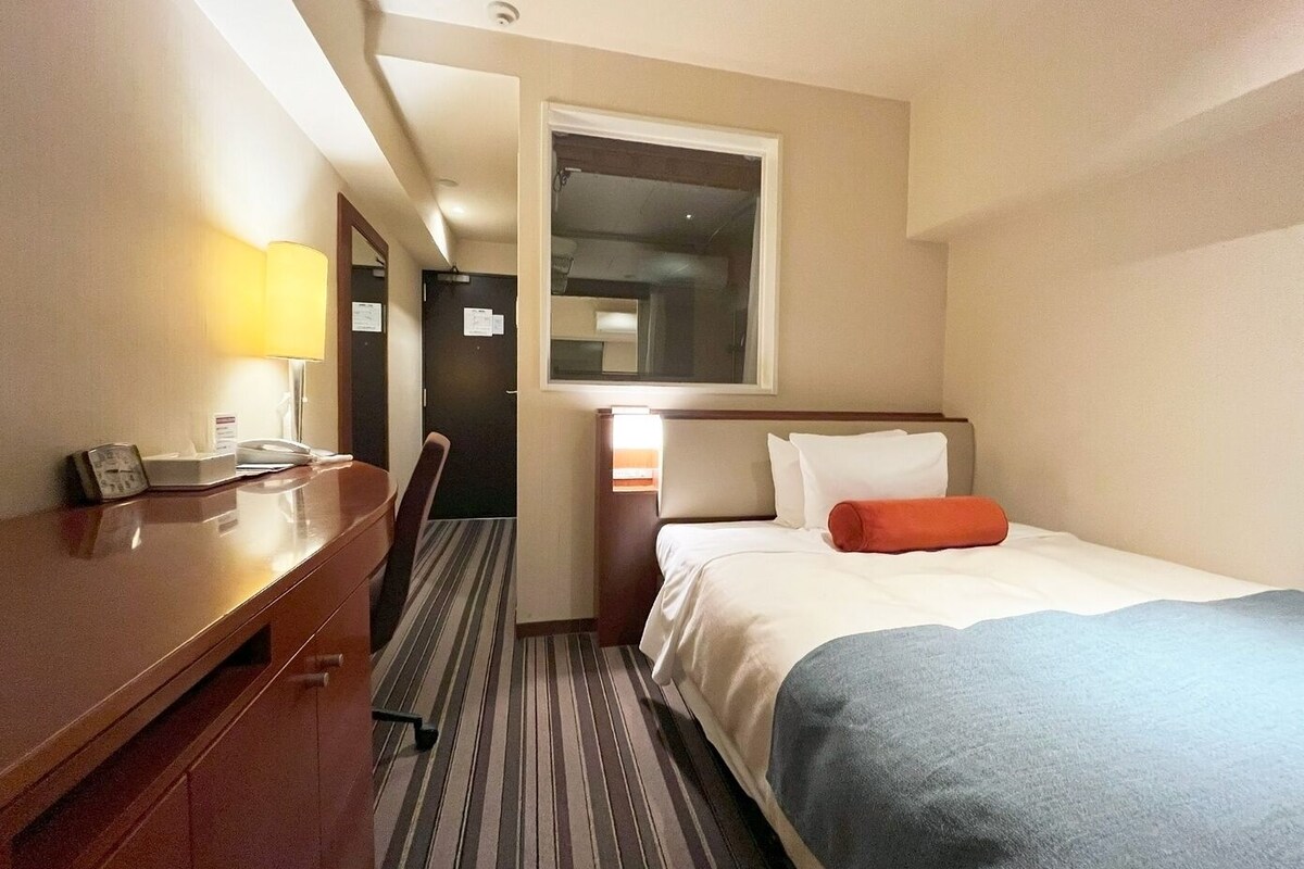 Ein Hotelzimmer mit Bett, Schreibtisch, Stuhl, Lampe und Fenster.