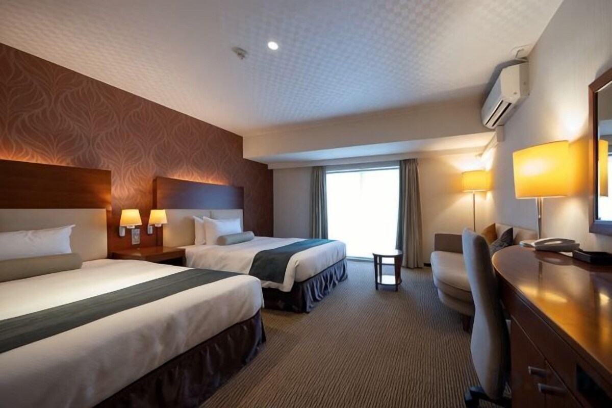 Ein Hotelzimmer mit zwei Betten, einem Schreibtisch, einem Stuhl, einem kleinen Tisch und einem Fenster mit Vorhängen.