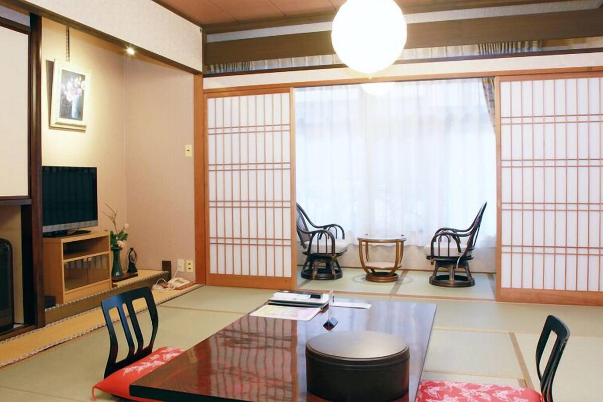 Um quarto japonês tradicional com tatames, uma mesa baixa, duas cadeiras, uma televisão e portas deslizantes.