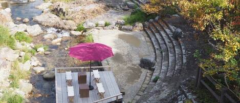 A wooden deck with a red umbrella, white chairs, and a table overlooking a river with a stone wall and autumn foliage.