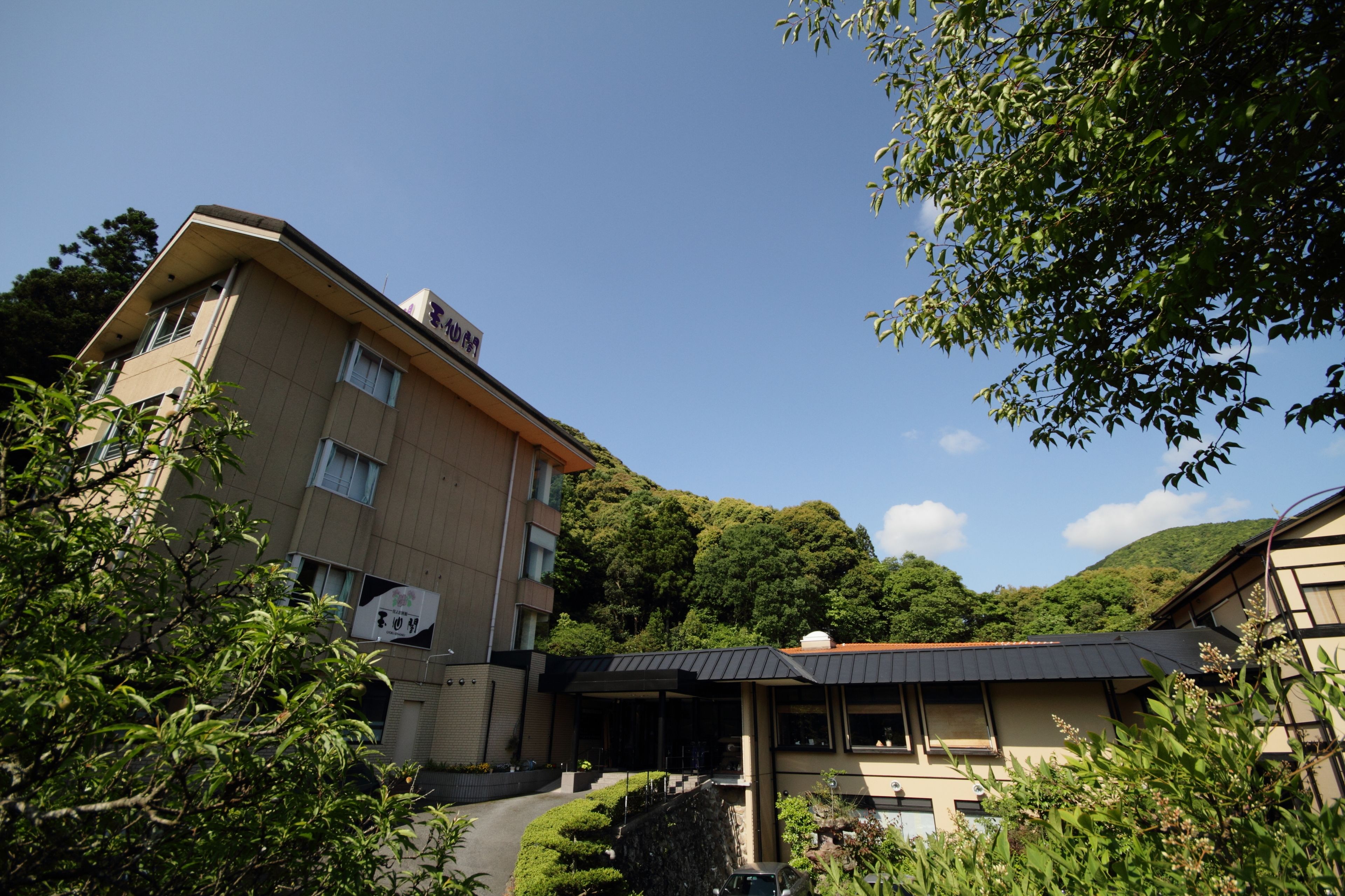 A multi-story building with a flat roof, surrounded by lush greenery and hills in the background.
