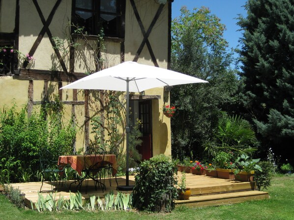 A patio with a table and chairs under a white umbrella, surrounded by potted plants and a wooden deck.