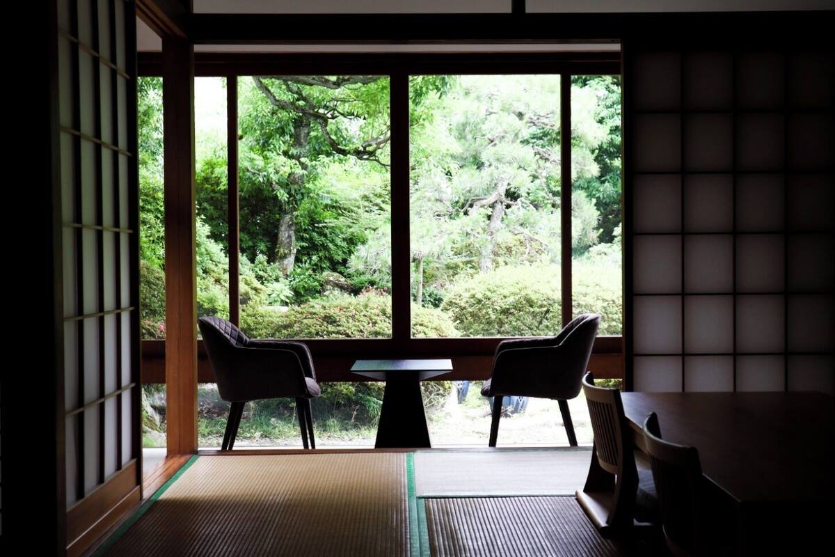 A traditional Japanese room with tatami flooring, a low table, two chairs, and a large window offering a view of a lush garden.