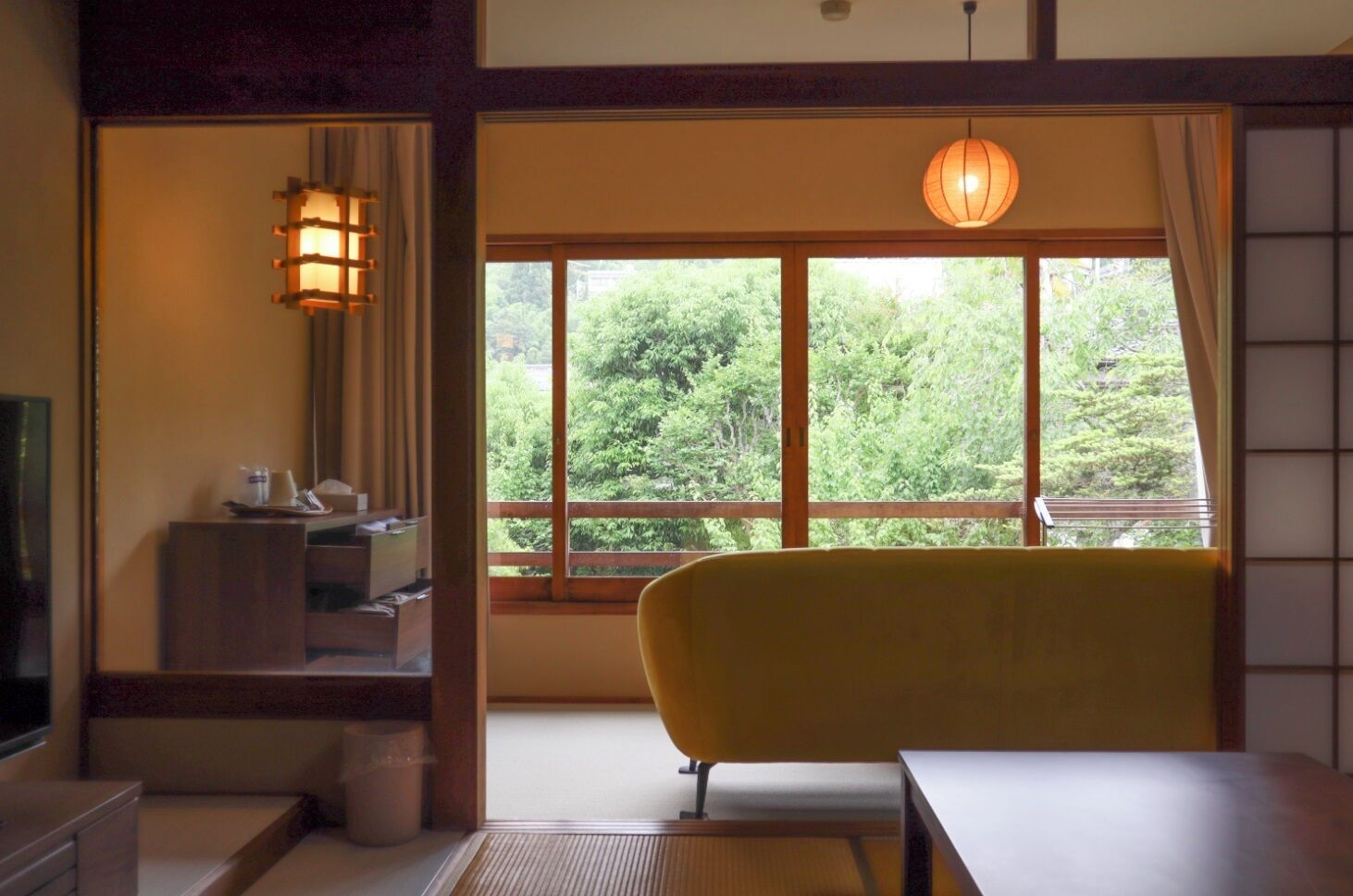 A room with a yellow chair, a table, a window with a view of greenery, a traditional Japanese lantern, and a small cabinet.