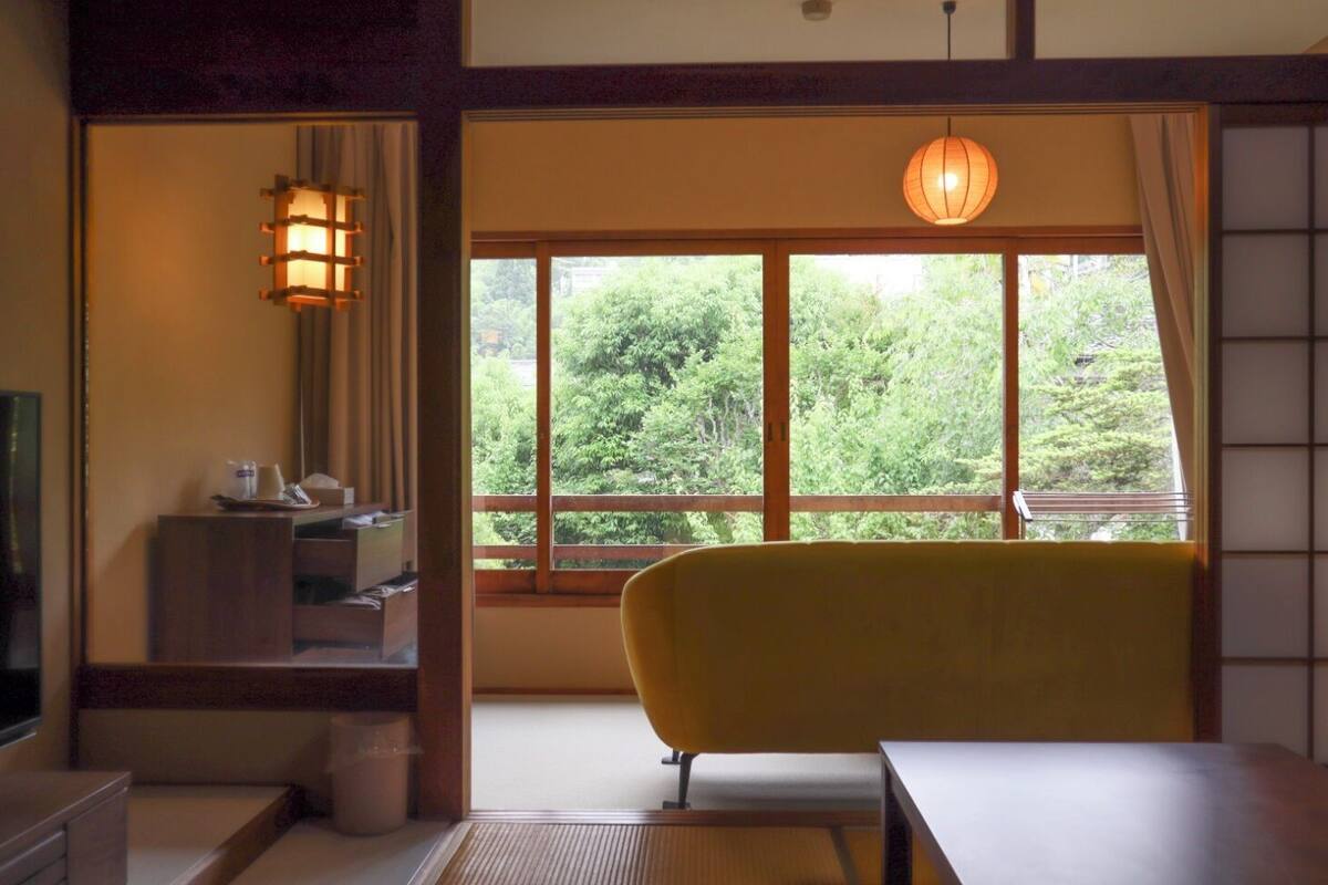 A room with a yellow chair, a table, a window with a view of greenery, a traditional Japanese lantern, and a small cabinet.