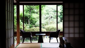 Ein traditioneller japanischer Raum mit Tatami-Bodenbelag, einem niedrigen Tisch, zwei Stühlen und einem großen Fenster mit Blick auf einen üppigen Garten.