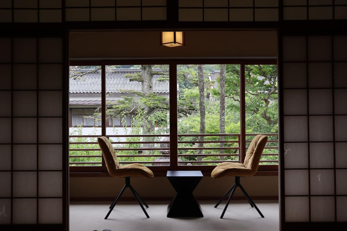 A traditional Japanese room with tatami flooring, a low table, two chairs, and a view of a traditional building and trees through a shoji screen.