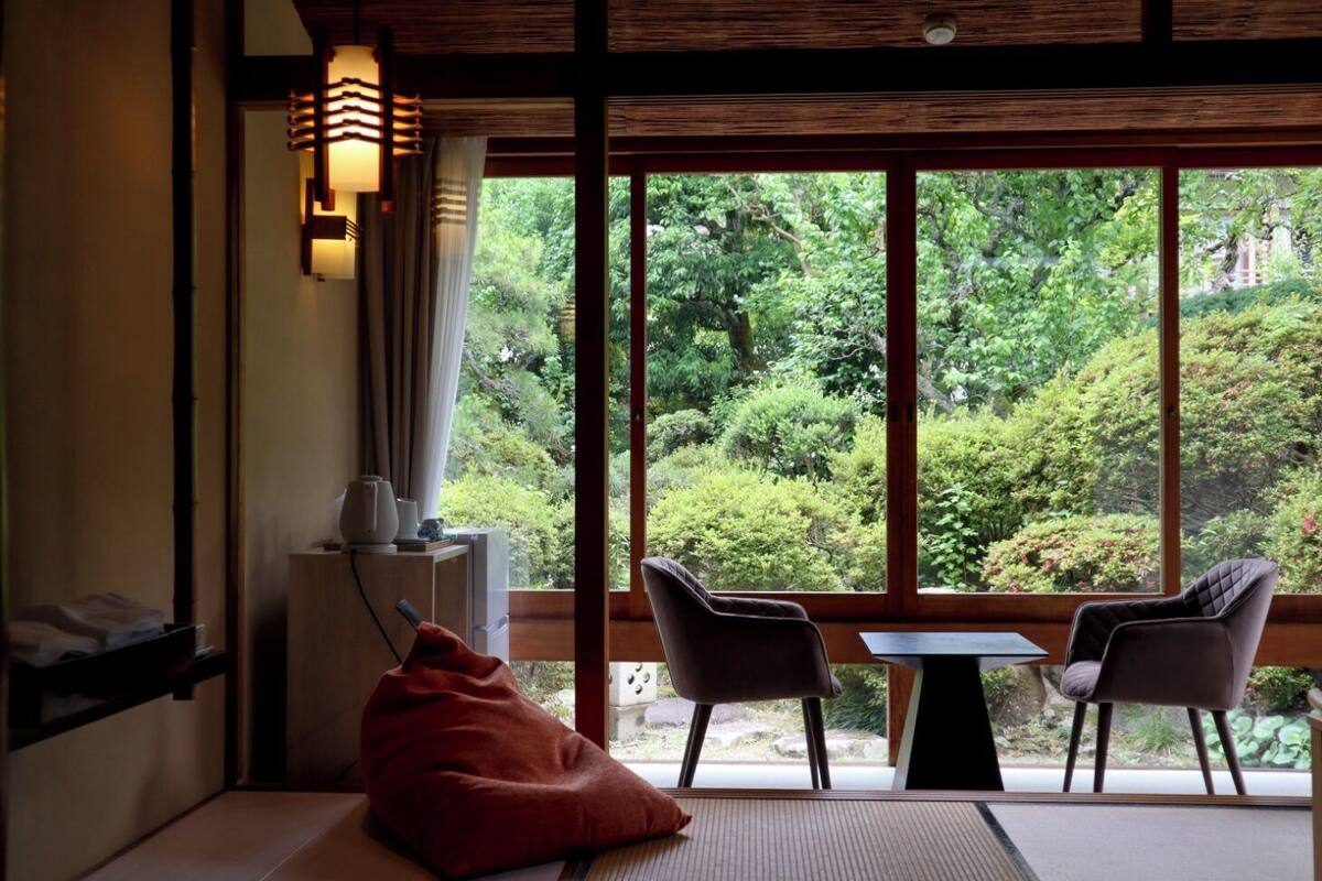 A room with tatami flooring, a low table, two chairs, a red bean bag, and a view of lush greenery through large windows.