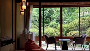Ein Zimmer mit Tatami-Boden, einem niedrigen Tisch, zwei Stühlen, einem roten Sitzsack und Blick auf üppiges Grün durch große Fenster.