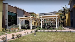 A courtyard with covered seating areas, a pathway, and a building with large windows and a palm tree. - Courtyard International Hotel (Lyantonde)
