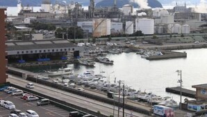 A marina with boats docked, a large industrial facility in the background, and a highway running parallel to the water.