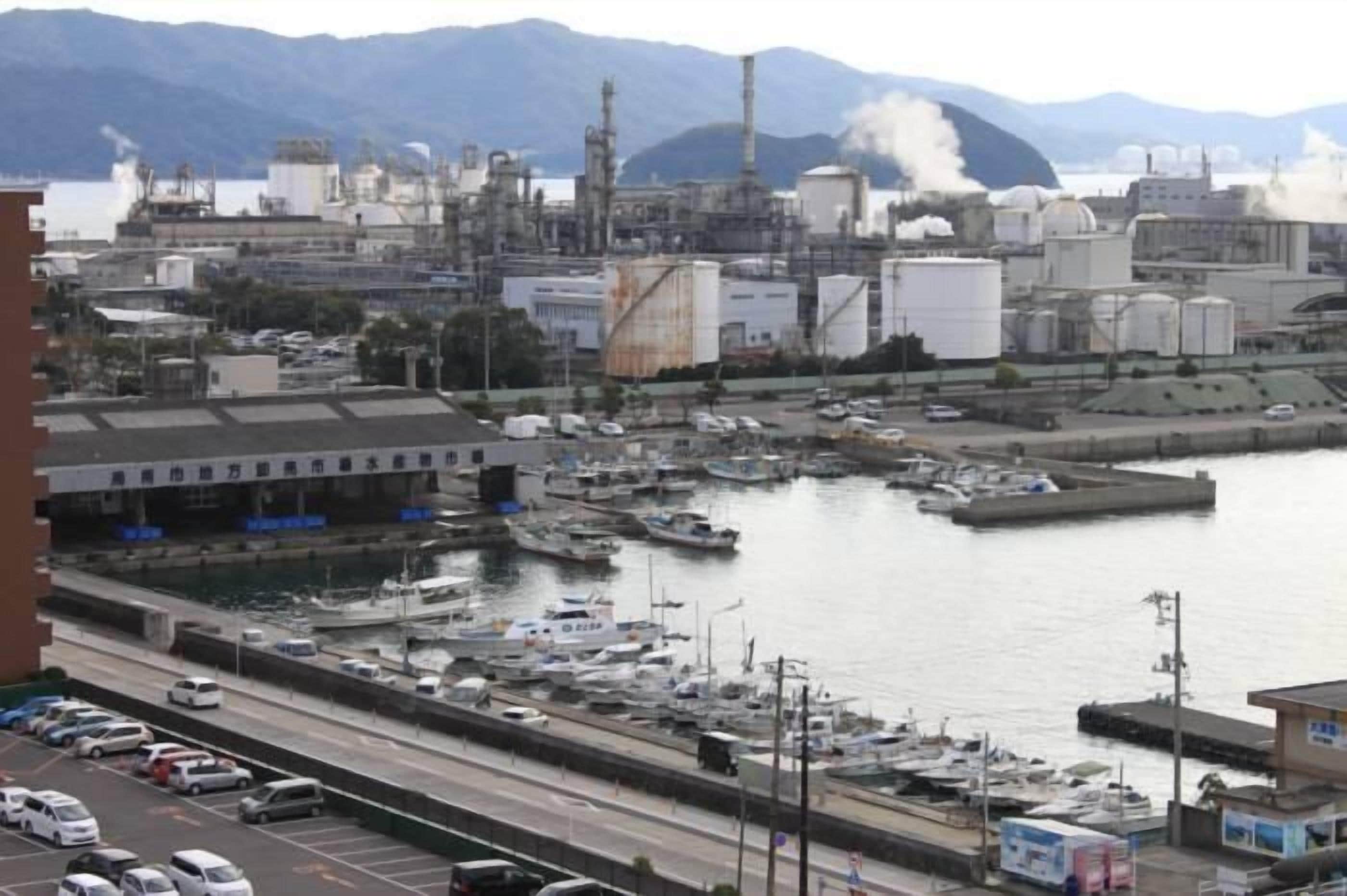 A marina with boats docked, a road with vehicles, and industrial buildings in the background.