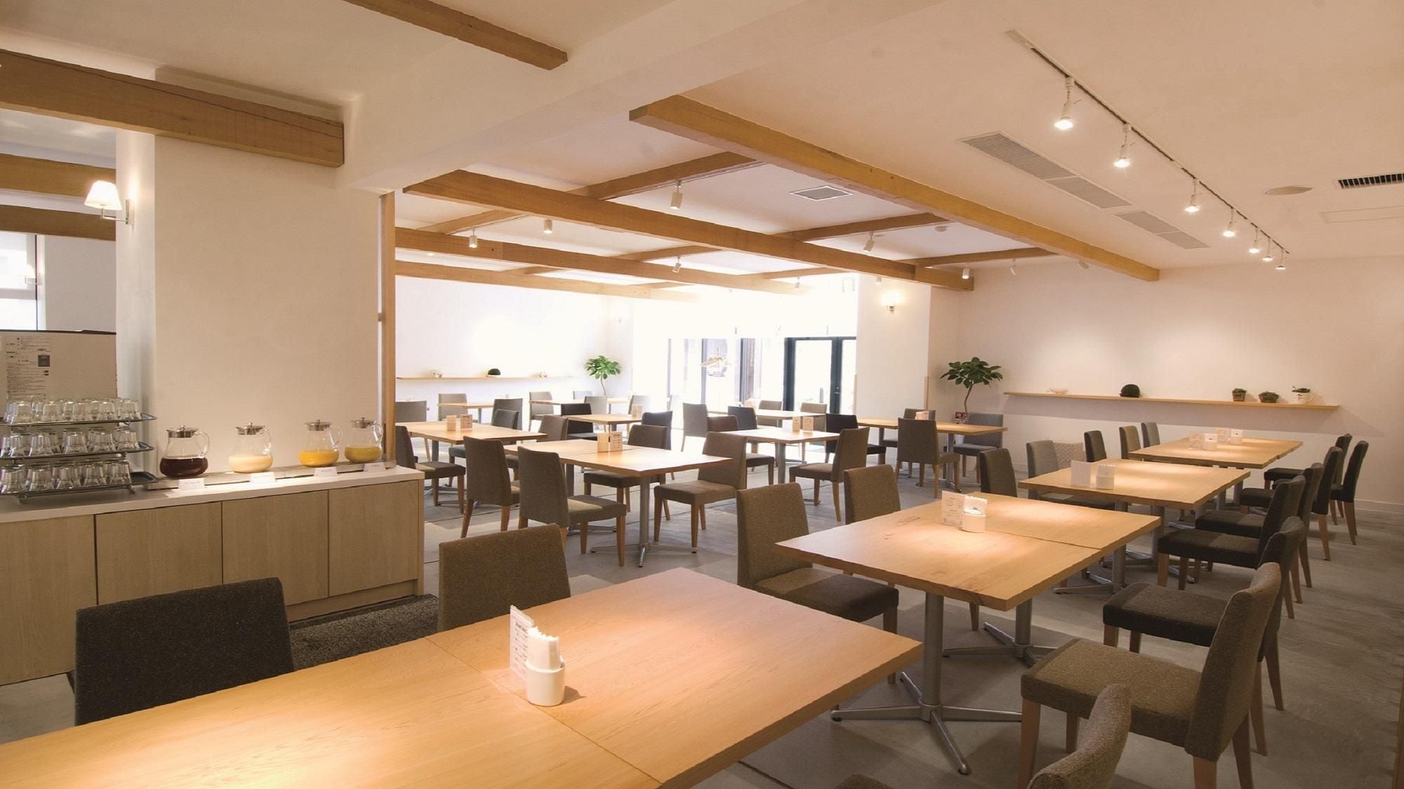 A spacious dining area with multiple tables and chairs, a kitchen area with a coffee station, and natural light from large windows.