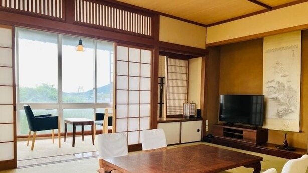 Een traditionele Japanse kamer met tatami vloerbedekking, een lage tafel, stoelen, een televisie en een schuifdeur naar een balkon.