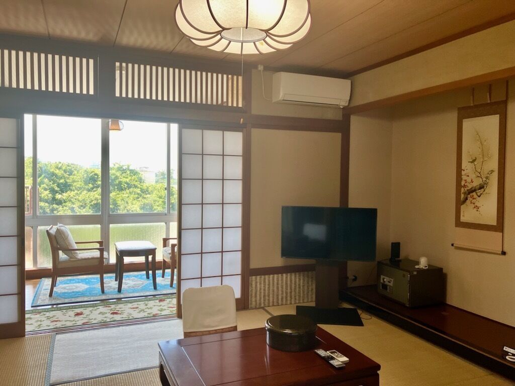 A traditional Japanese-style room with tatami flooring, a television, a sofa, and a sliding door leading to a balcony.