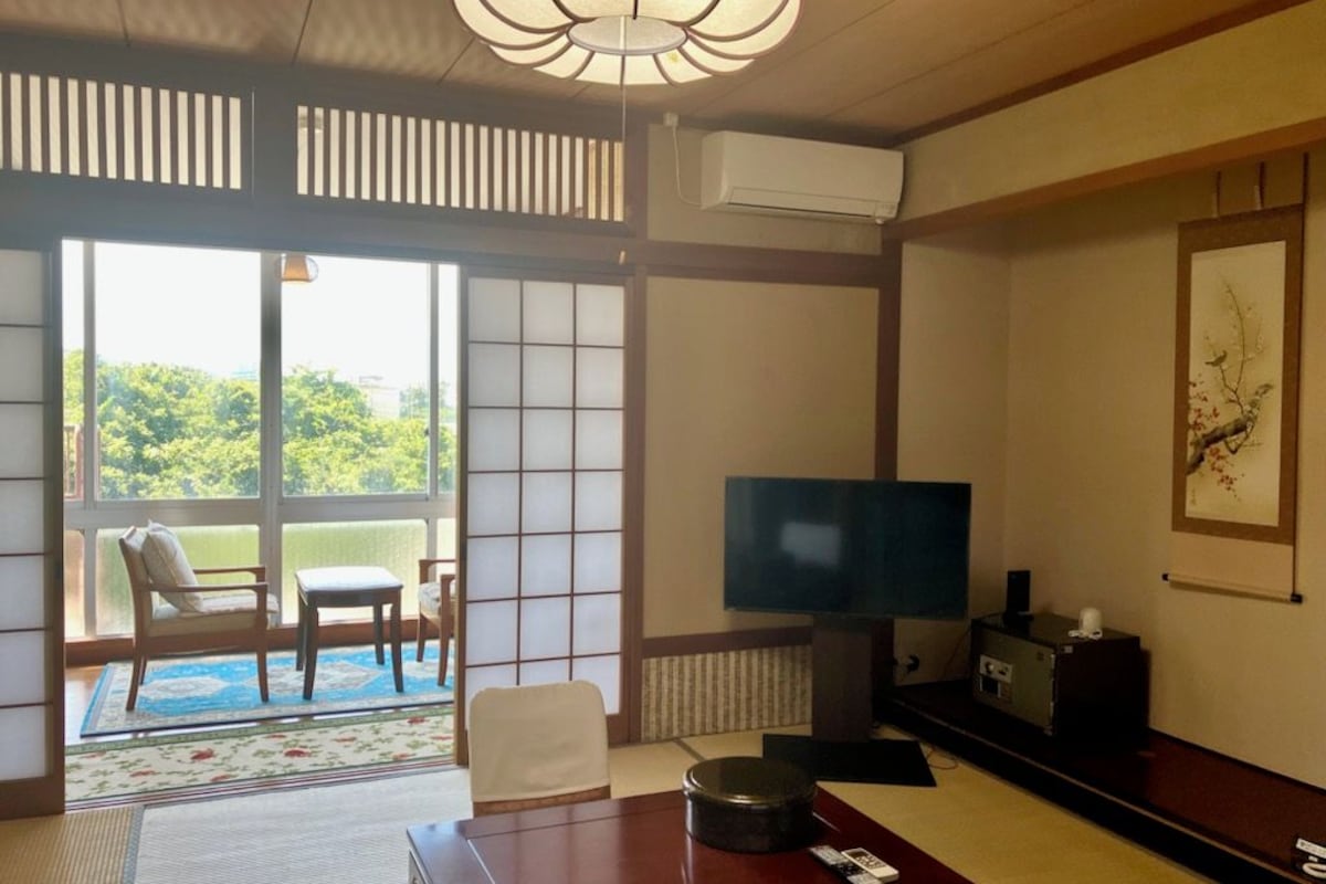 A traditional Japanese-style room with tatami flooring, a television, a sofa, and a view of greenery outside.