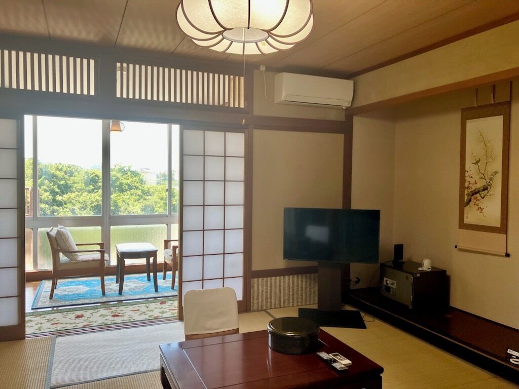 A traditional Japanese-style room with tatami flooring, a television, a sofa, and a view of greenery outside.