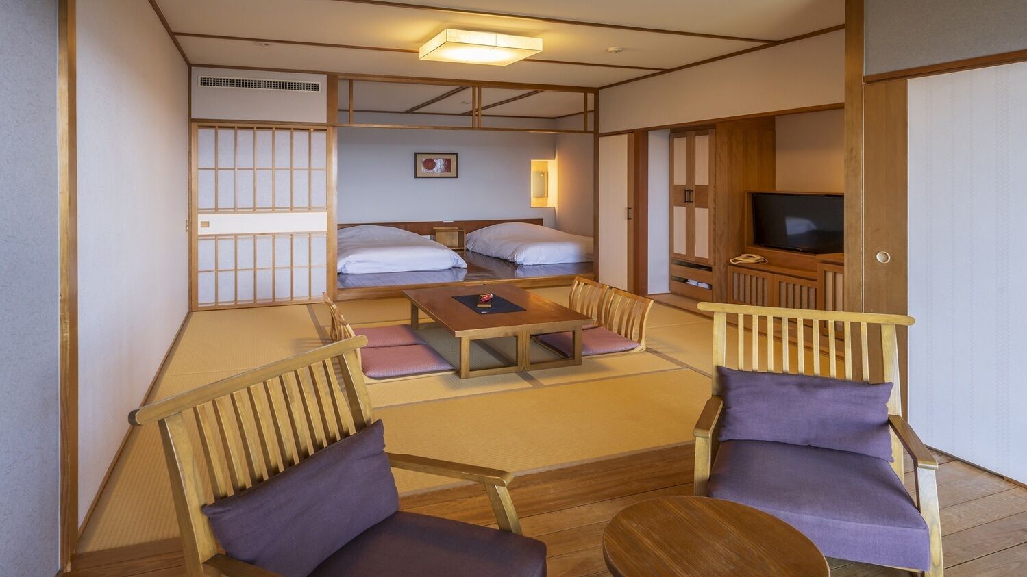 Ein traditioneller japanischer Raum mit Tatami-Böden, einem niedrigen Tisch, zwei Tatami-Matten, einem Fernseher und einer Schiebetür.