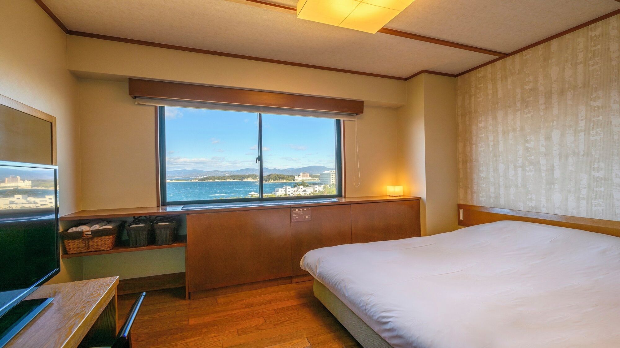 Ein Hotelzimmer mit Bett, Fernseher, Fenster mit Meerblick und einem Holztisch mit Körben.